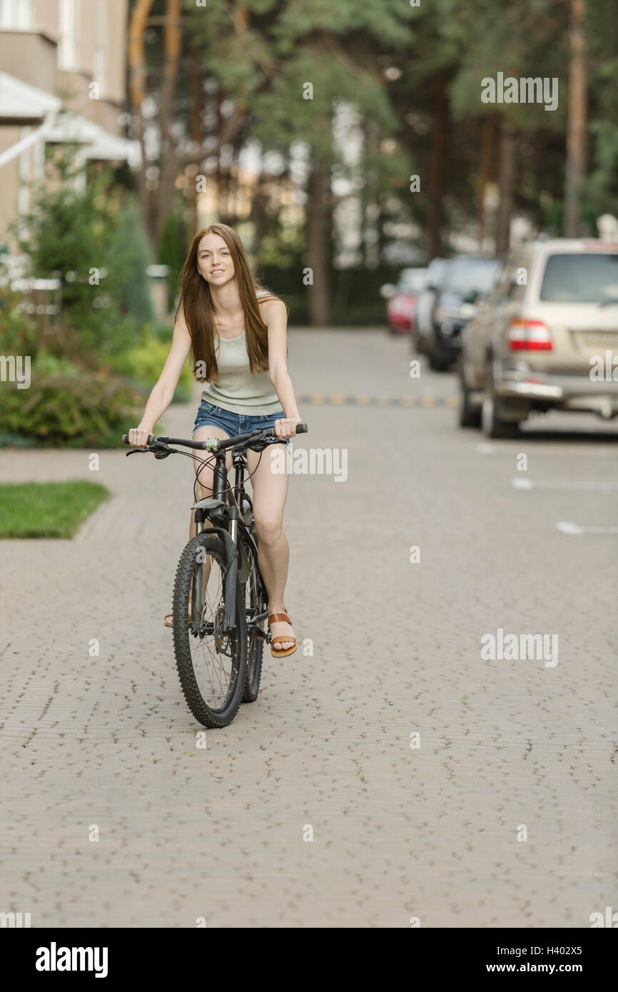 Lächelnde Frau Radfahren auf gepflasterten Straße gegen geparkte Autos in der Stadt Stockfoto