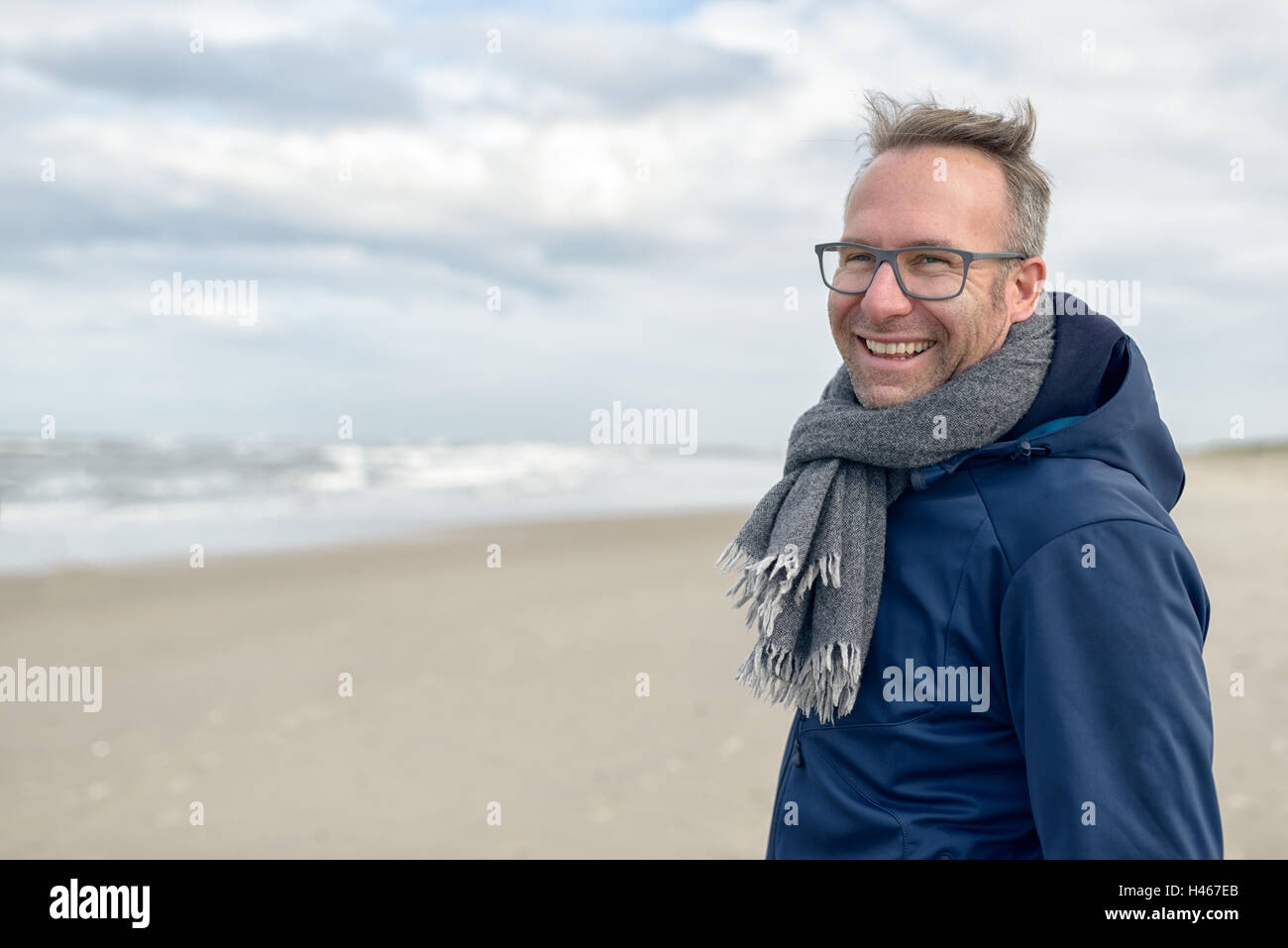 Lächelnder Mann mittleren Alters tragen Brillen und einen Schal aus Wolle Stand an einem einsamen Strand im Herbst an einem bewölkten Tag mit cop Stockfoto