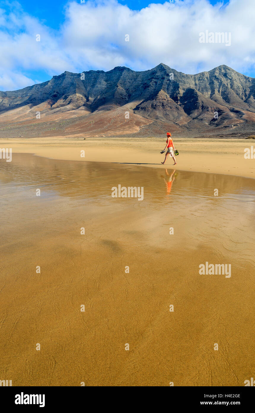 Junge Frau in orange Hemd und Hut gehen auf nassen Sand St Cofete Strand mit Bergen im Hintergrund, Fuerteventura, Kanarische Inseln Stockfoto