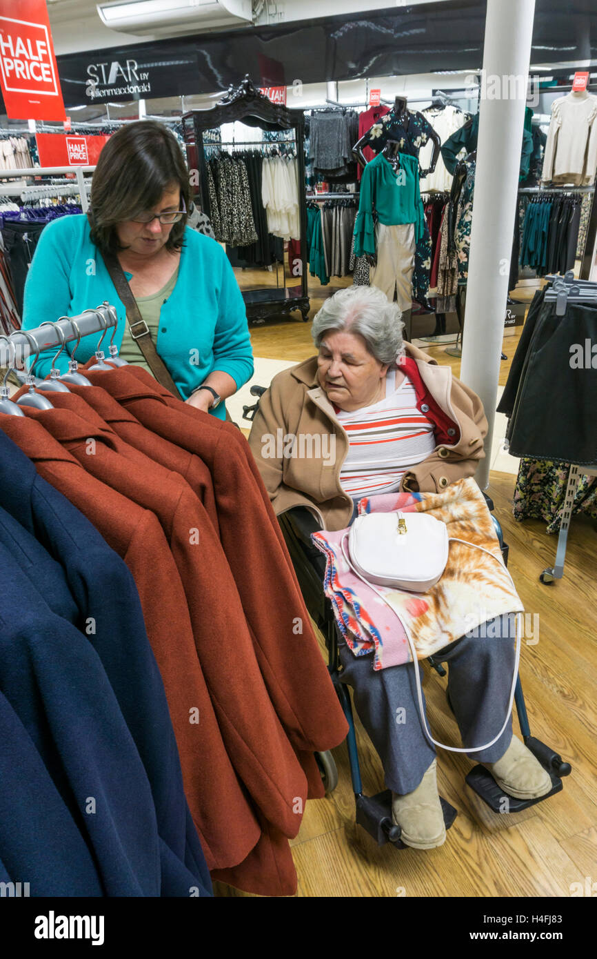 Eine ältere Dame im Rollstuhl Geschäfte für eine neue Jacke in einem Kaufhaus mit Ihrem Betreuer oder Helfer. Stockfoto