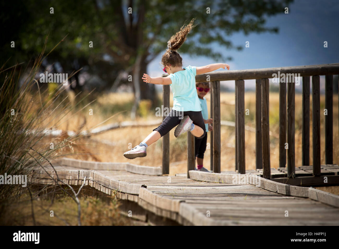 Rückseite des kleinen Mädchens Schwanz springen in der Luft über dem hölzernen Gehweg mit Armen erweitert. Ihre Schwester stehend auf Holz Stockfoto