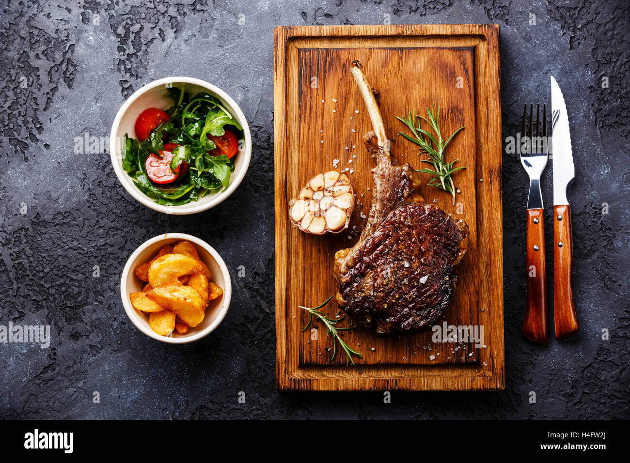 Vom Grill Grill Steak auf Knochen Kalbfleisch Rippe mit Kartoffelspalten und Salat mit Tomaten und Rucola auf dunklem Hintergrund Stockfoto