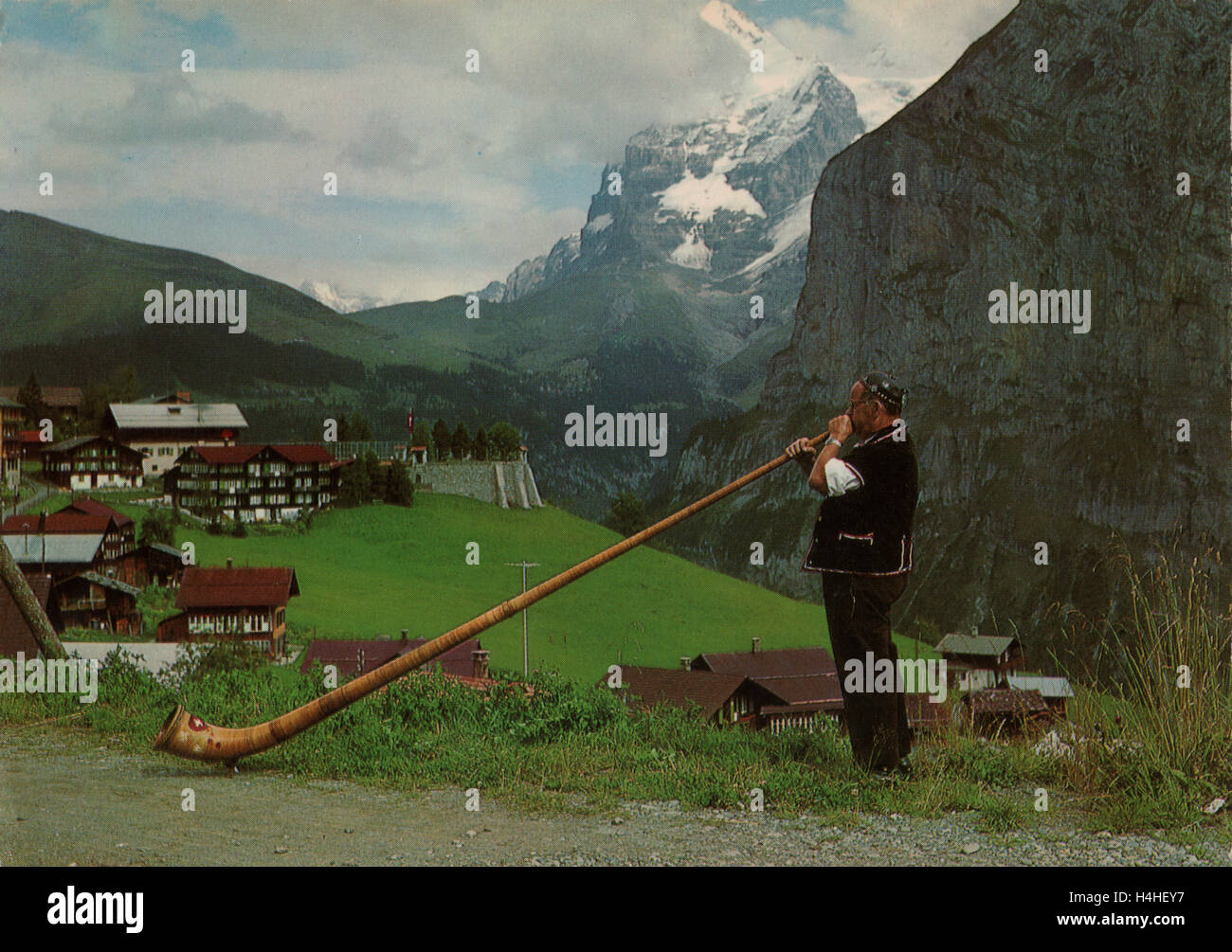 Alte Ansichtskarte eines Mannes in Schweizer Trachtenmode weht ein Alphorn oder Alphorn mit einer Schweizer Stil Berg- und ländlichen Hintergrund ca. 1960er Jahre. Stockfoto