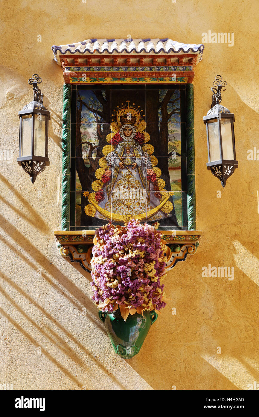 Spanische Jungfrau (Virgen del Rocio) Fliese auf eine gelbe Wand mit Laternen auf beiden Seiten und einen Topf mit Blumen unten Stockfoto