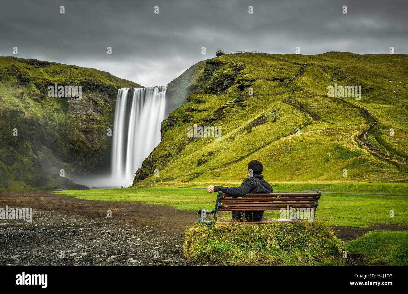 Wanderer auf einer Bank sitzt und schaut auf den berühmten Skogafoss Wasserfall im Süden Islands Stockfoto