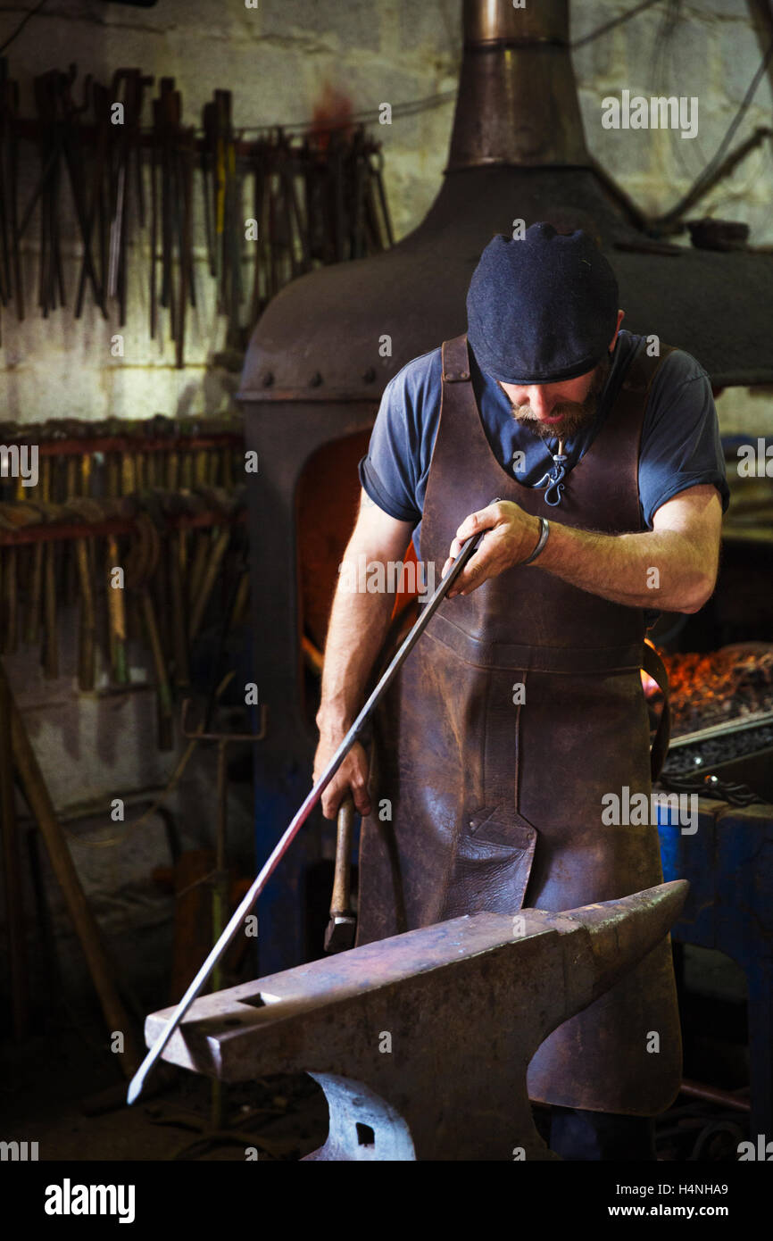 Ein Schmied in eine Lederschürze hält eine dünne Länge aus Metall gegen Amboss in einer Werkstatt. Stockfoto
