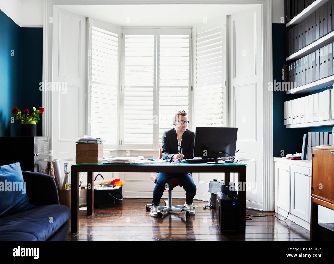 Ein Mann in Anzug und Trainer mit Brille, sitzen an einem Schreibtisch in einem Erker. Eine leichte luftige Büro mit Dateien in den Regalen. Stockfoto
