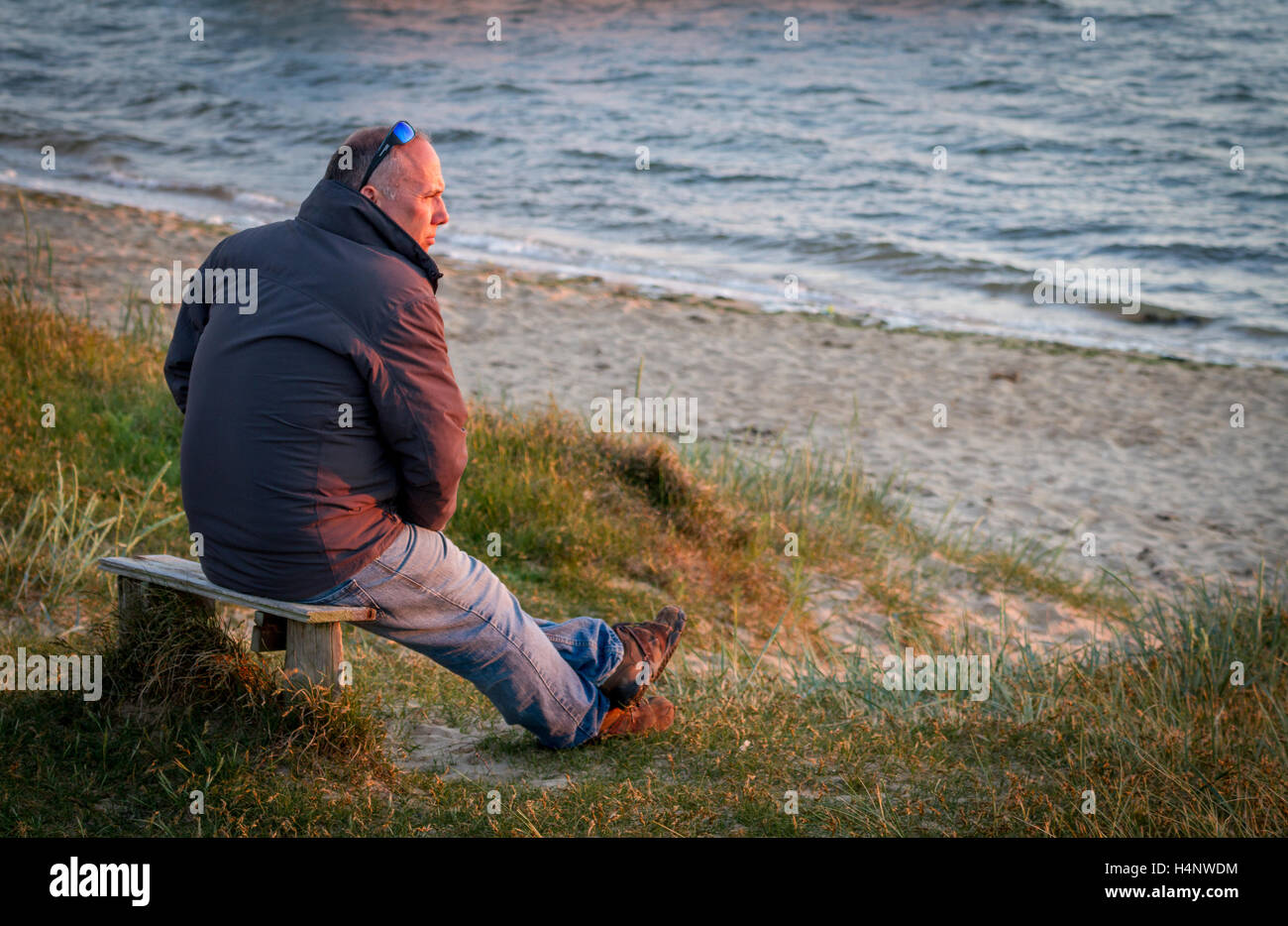 Mann sitzt auf Sitzbank mit Jacke und Sonnenbrille genießen den Blick auf den Sonnenuntergang über der Bucht Stockfoto