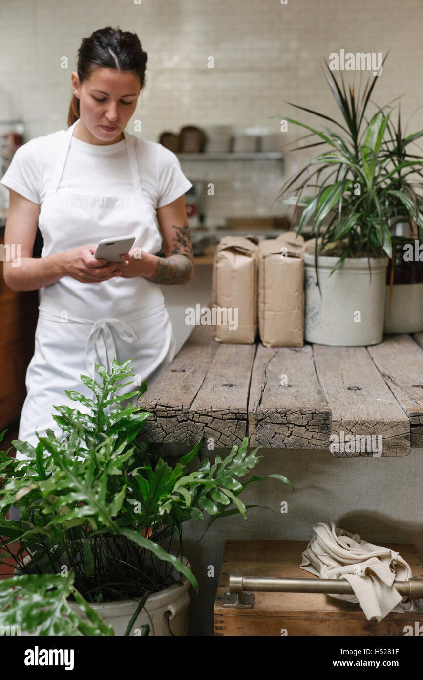 Frau trägt eine weiße Schürze in einer Küche mit einem Mobiltelefon. Stockfoto