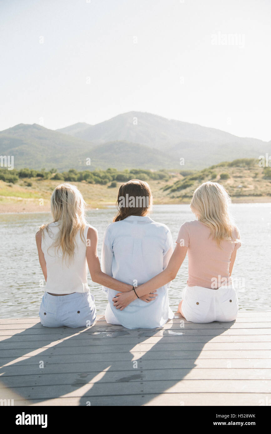 Rückansicht einer Mutter und ihren beiden blonden Töchtern sitzen auf einem Steg. Stockfoto
