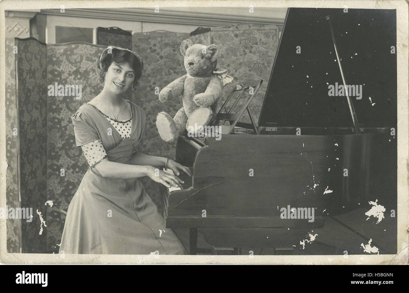 Eine alte Fotopostkarte mit einer Frau, möglicherweise Schauspielerin Marjorie Neil, und einem Spielzeugbären, der am Klavier sitzt und einen nostalgischen Moment aus der Unterhaltung des frühen 20. Jahrhunderts festnimmt. Stockfoto