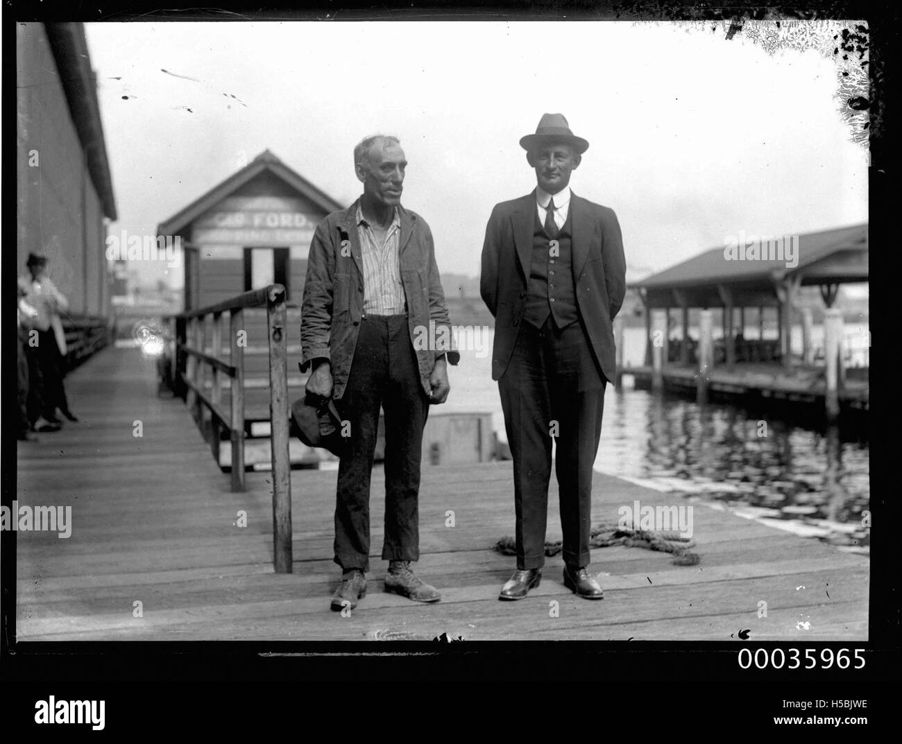 Dieses Foto aus der KAPUZENKOLLEKTION zeigt zwei Männer, die zwischen 1920 und 1953 auf einem Kai in Sydney standen. Das Bild fängt die industrielle und maritime Atmosphäre der damaligen Zeit ein, mit Gebäuden und Festivals, die die nationale Szene widerspiegeln. Stockfoto