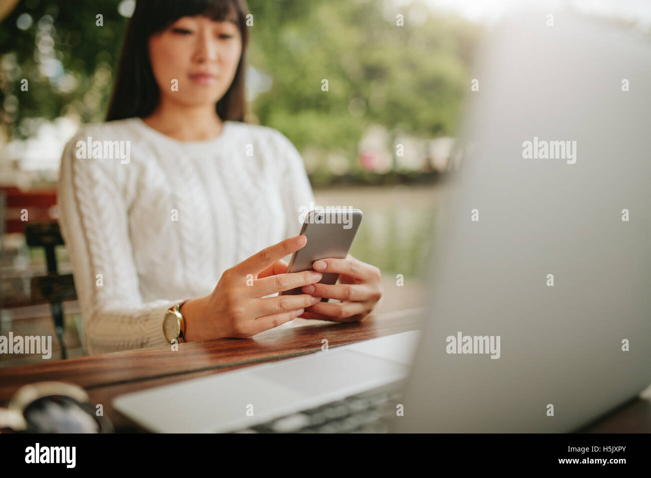 Schuss von Asiatin mit Handy in Straßencafé. Junge Frau mit Laptop lesen SMS-Nachricht auf ihr ce am Tisch sitzen Stockfoto