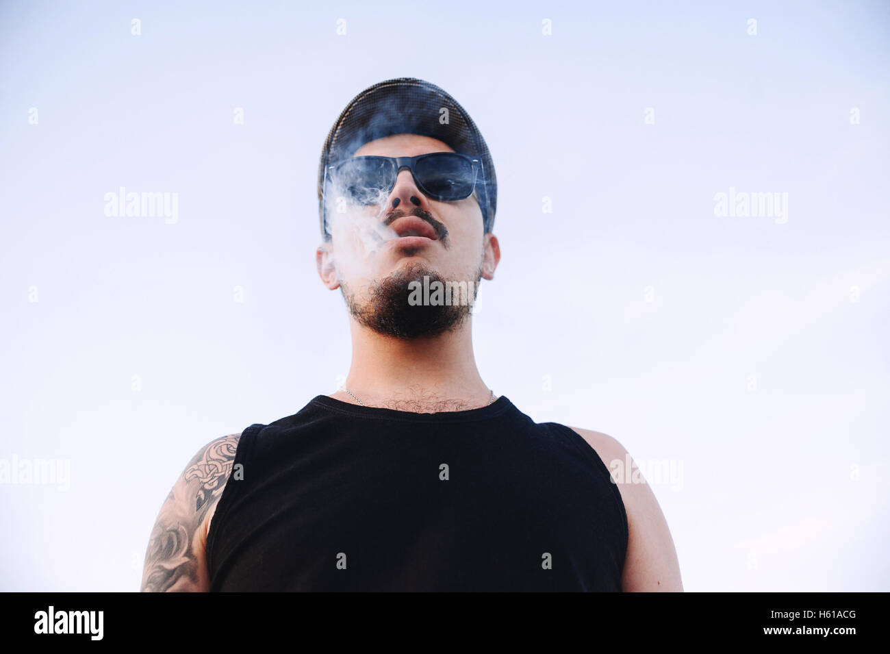 Nahaufnahme, Porträt des jungen Kerl Sonnenbrille eine Mütze mit Bart und Schnurrbart mit Tätowierungen, Ausatmen Rauch Zigarette Stockfoto