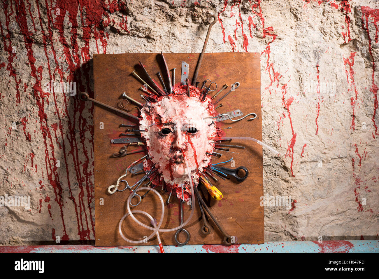 Dunkelhäutigen blutiges Gesicht einer Person auf einem Holzbrett mit sortierten scharfen Waffen zusammen mit Blut bespritzt Wand in einem Halloween-Horror-Konzept gedehnt geöffnet. Stockfoto