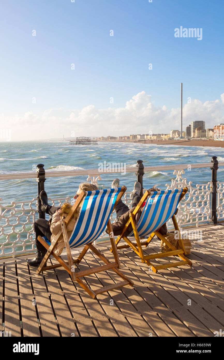 Zwei Personen am Meer sitzen in Liegestühlen in der Sonne am Pier von Brighton, Brighton, East Sussex England UK Stockfoto
