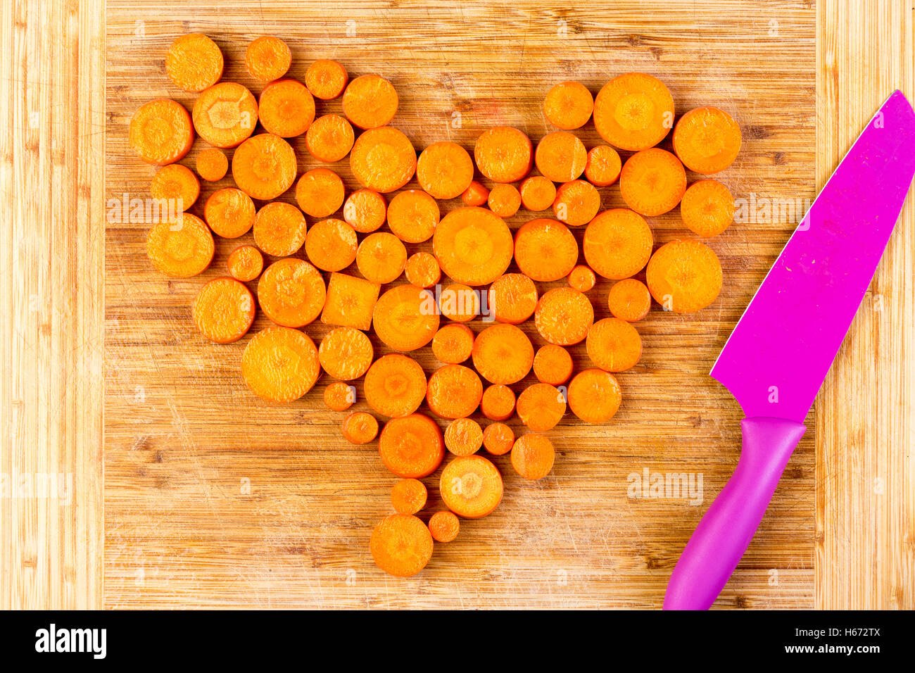 Top-down Ansicht auf verschiedenen großen gehackte rohe orange farbigen Karotten angeordnet in Herzform neben lila Messer auf Holz schneiden Stockfoto