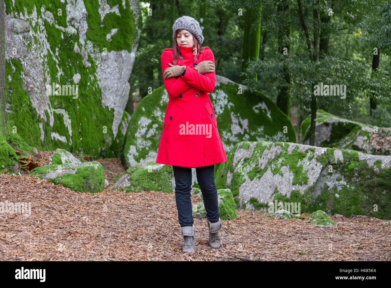 Junge Frau vor Kälte zitternd und umarmen sich auf einen Wald tragen einen roten Mantel, eine Mütze und Handschuhe im winter Stockfoto