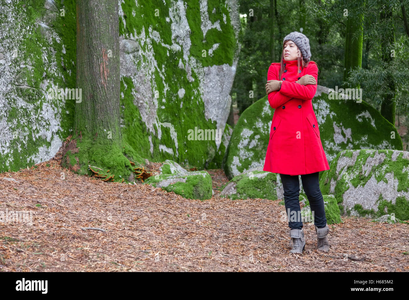 Junge Frau vor Kälte zitternd und umarmen sich auf einen Wald tragen einen roten Mantel, eine Mütze und Handschuhe im winter Stockfoto
