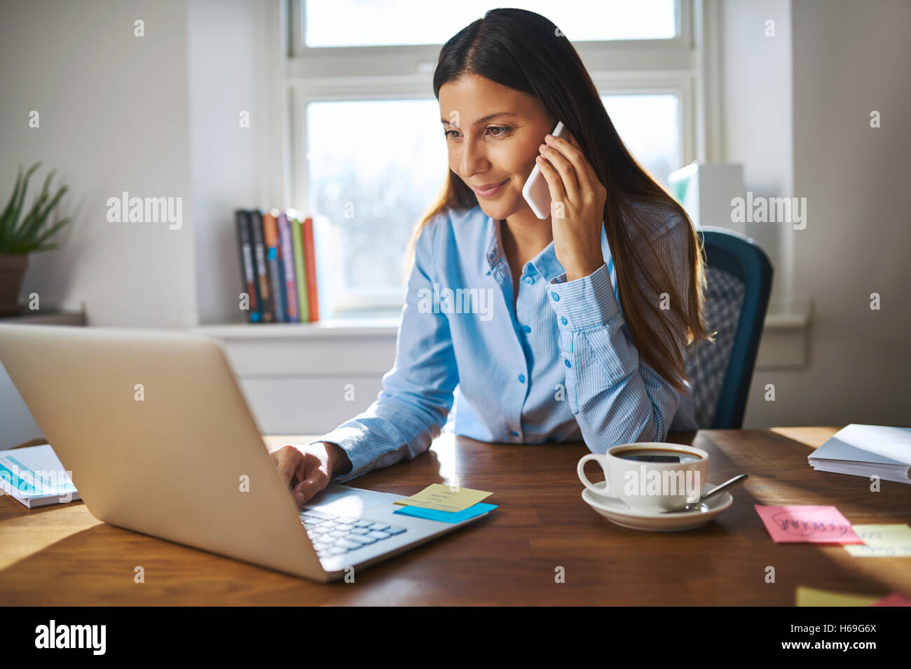 Süße junge weibliche Erwachsene mit Grinsen auf Handy während der Arbeit am Laptop-Computer am Schreibtisch neben Kaffeetasse mit großen Fenster auf der Rückseite Stockfoto