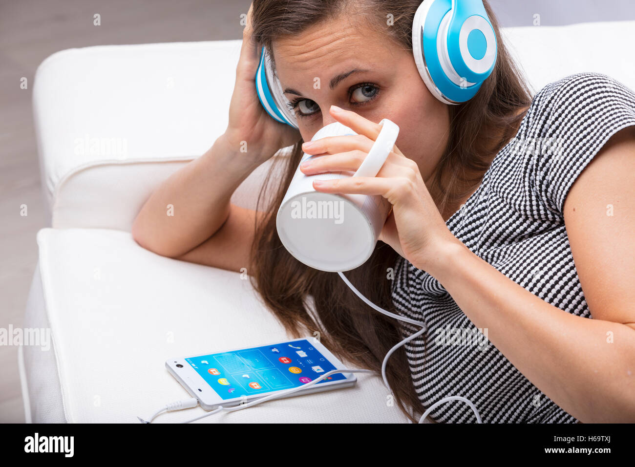 Porträt einer jungen Frau, das Anhören von Musik über Kopfhörer über ihr Smartphone beim Trinken etwas von ihrem Becher und watchi Stockfoto
