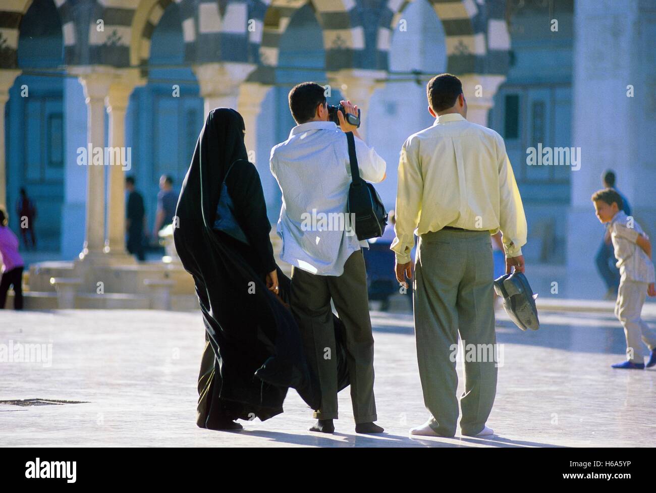 Eine Gruppe von Besuchern, die Umayyaden-Moschee in Damaskus in Damaskus, zwei Männer und eine verschleierte Frau, so dass ein Film ihres Besuchs. Aufgenommen am 21.08.2001. Man sieht deutlich aus diesem Bild, das es nicht erlaubt ist, geben eine Moschee mit Schuhen, die auf der Straße getragen worden sein. Die Umayyaden-Moschee in Damaskus ist eine der ältesten der Welt. Es wurde im Stil einer alten Basilika erbaut und in vorislamischer Zeit war die Kathedrale, wo Johannes der Täufer gewidmet war. Traditionelle Aufzeichnungen, zufolge wird seinen Kopf in einem Schrein beibehalten. Die Moschee enthält einen umfangreiche Hof mit drei kleineren Stockfoto