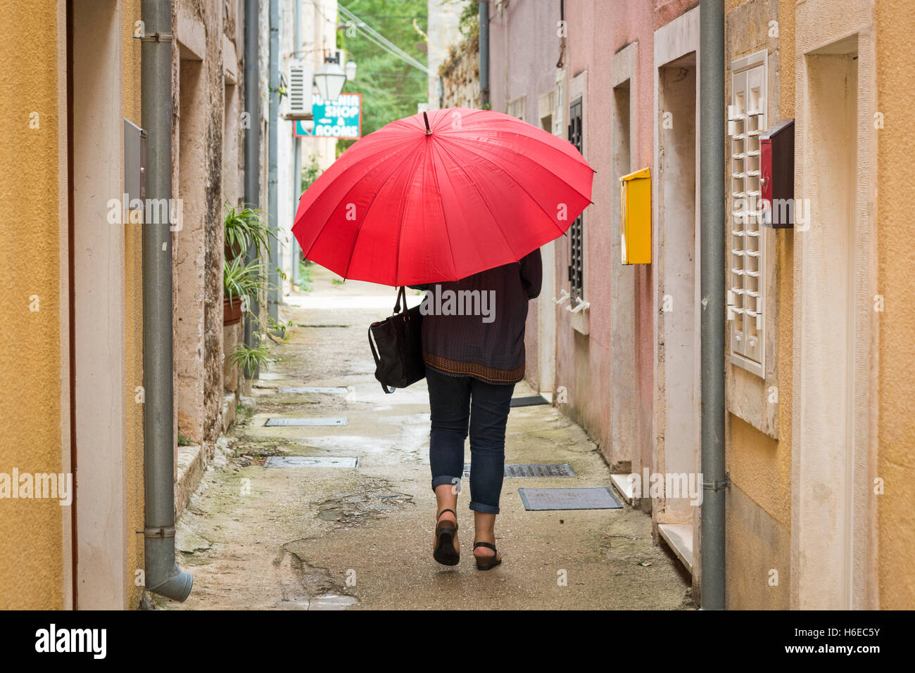 Eine Frau, die einen leuchtenden roten Regenschirm durch eine schmale Gasse in Ston Kroatien an einem bewölkten und regnerischen Tag Stockfoto