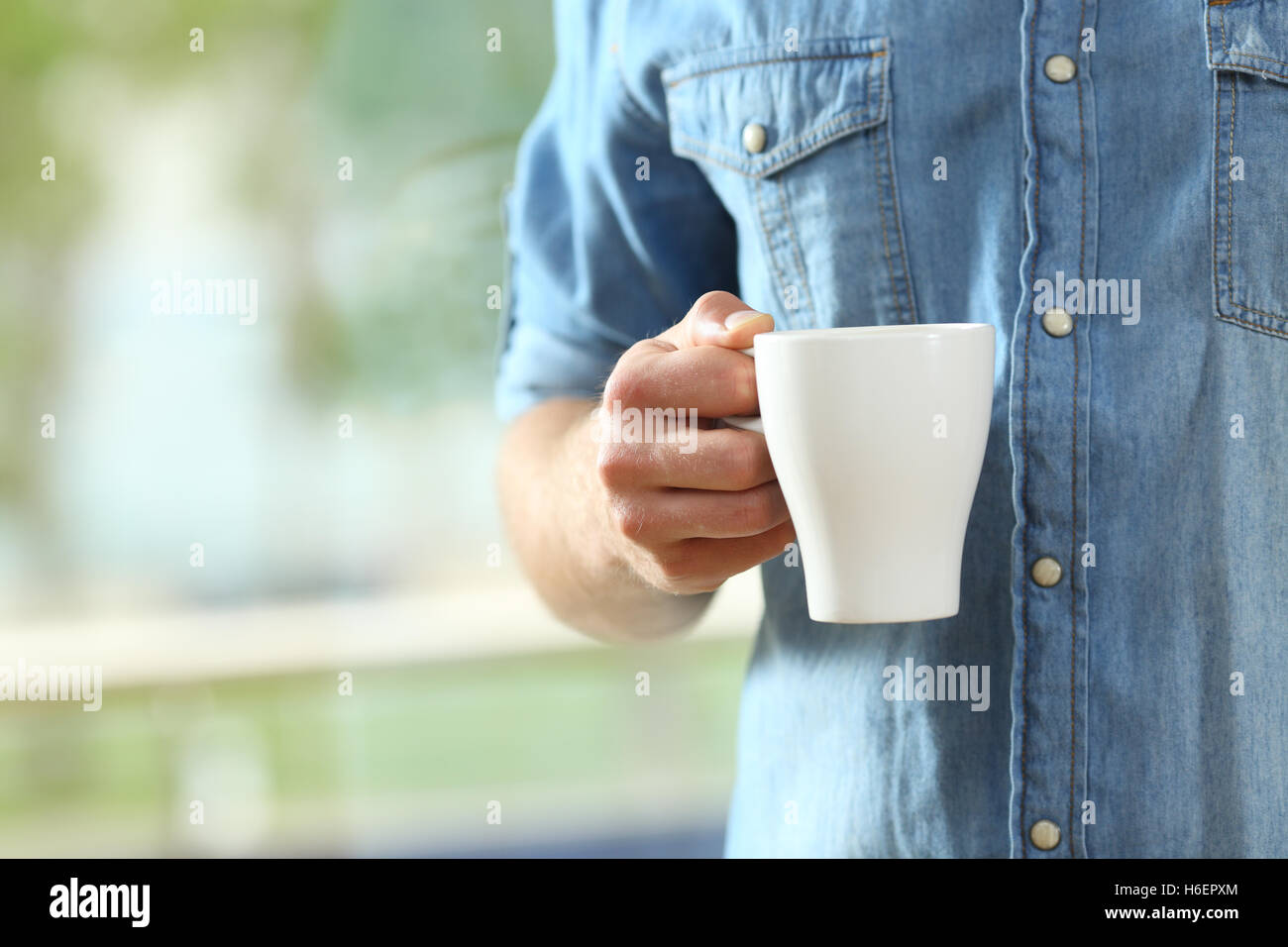 Nahaufnahme von einer Hand des Mannes hält eine Kaffeetasse neben ein Fenster mit einem grünen Hintergrund außerhalb Stockfoto