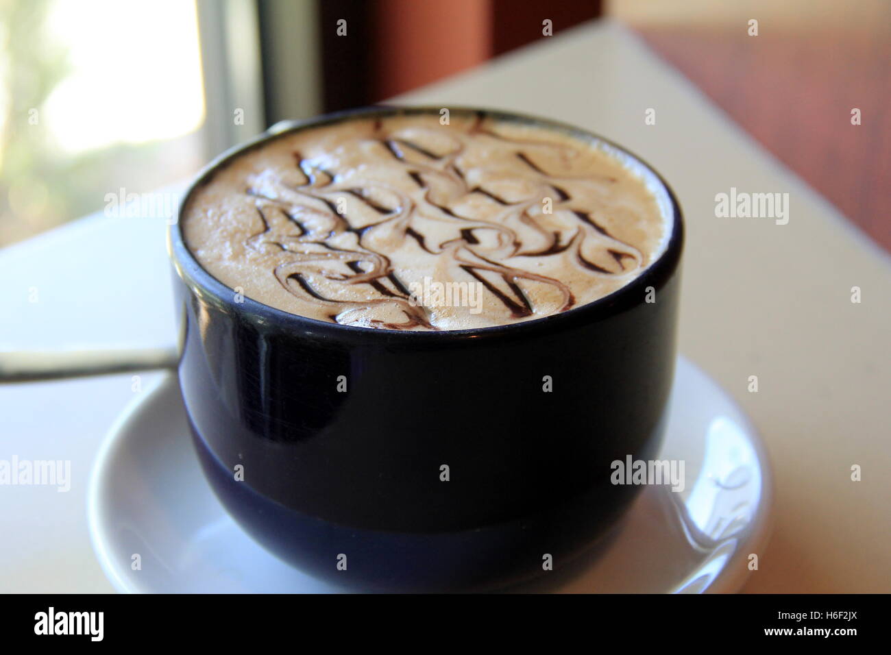 Köstlicher Mocha Cappuccino in tiefblauer Tasse auf weißer Untertasse in der Nähe von Fenster im Restaurant, ein herzlicher Empfang für Gäste. Stockfoto