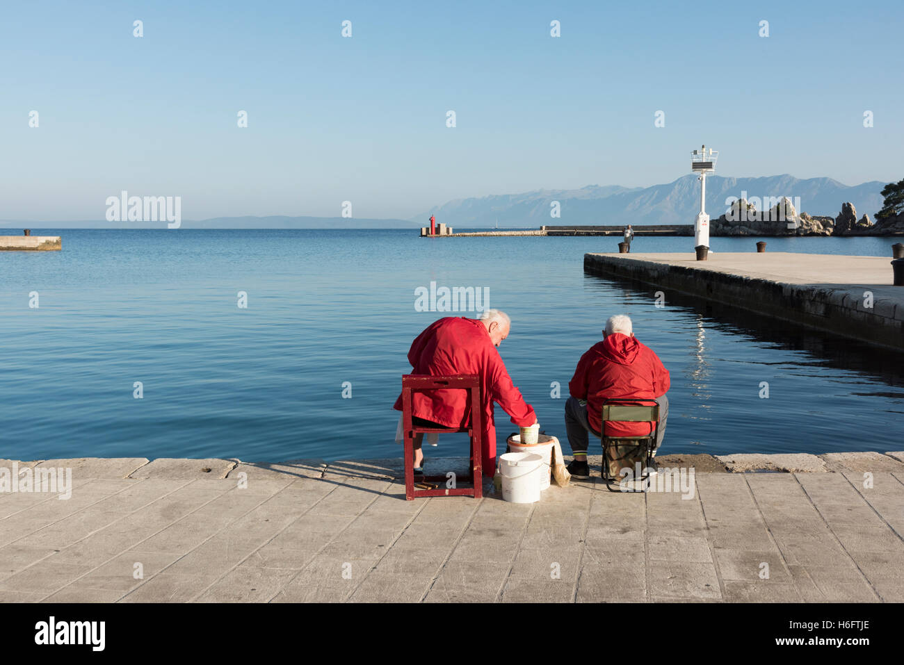 Zwei ältere Männer tragen rote Mäntel sitzen auf der Hafenmauer in Trpanj Kroatien Angeln im Meer Stockfoto