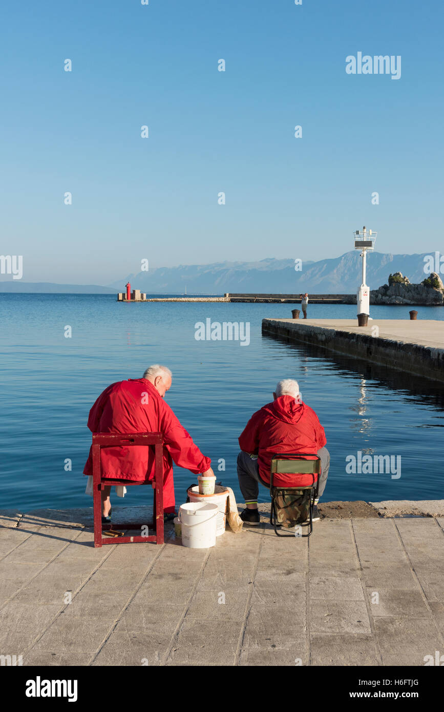 Zwei ältere Männer tragen rote Mäntel sitzen auf der Hafenmauer in Trpanj Kroatien Angeln im Meer Stockfoto