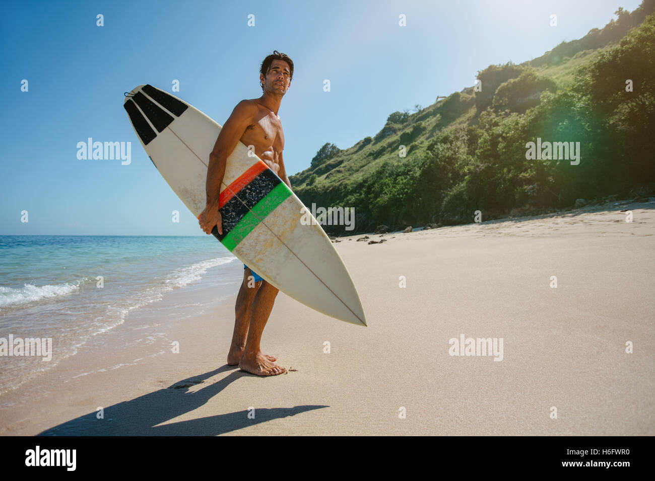 Voller Länge Schuss des jungen Mannes mit Surfbrett am Strand. Männliche Surfer Surfbrett am Ufer Meeres halten und wegsehen. Stockfoto