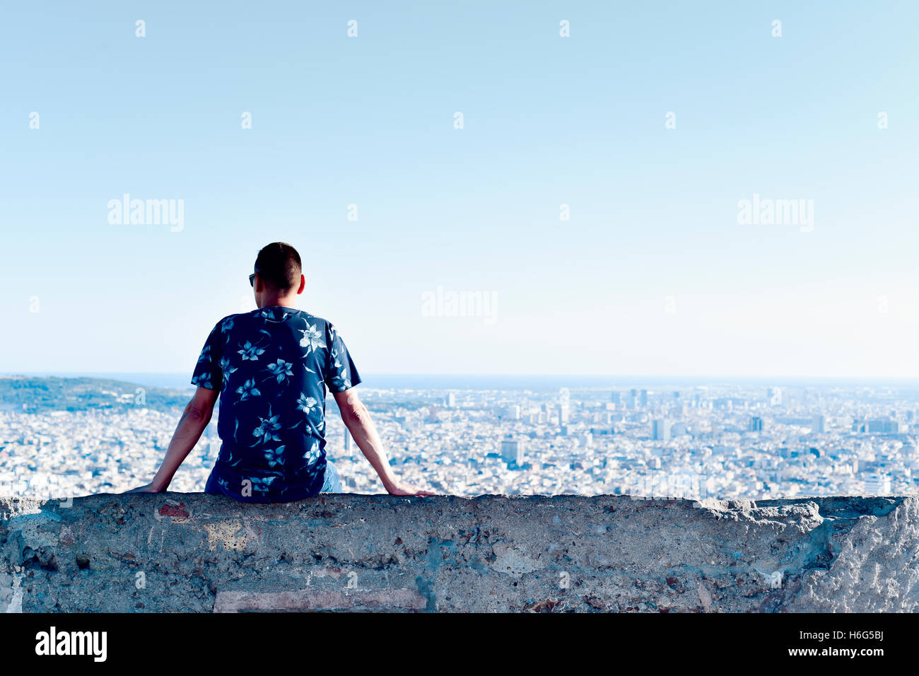 ein junger kaukasischen Mann, von hinten gesehen sitzt an der Spitze eines Hügels, Beobachtung der Stadt unter ihm Stockfoto