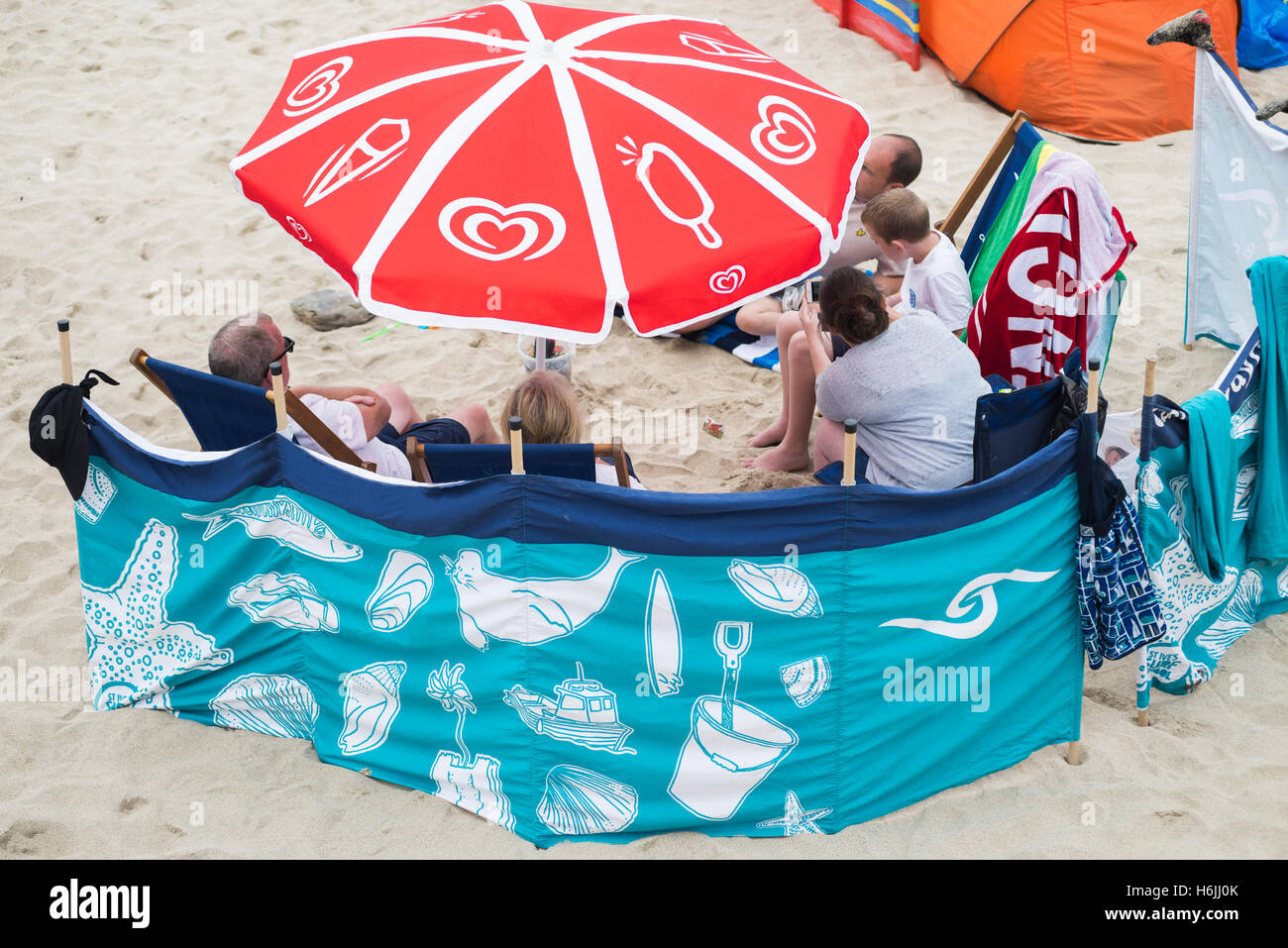 Familie als Urlauber am Strand von St Ives sitzen und reden hinter einem bunten Windschutz und Sonnenschirm an einem bewölkt und windig Sommertag, Cornwall, UK Stockfoto
