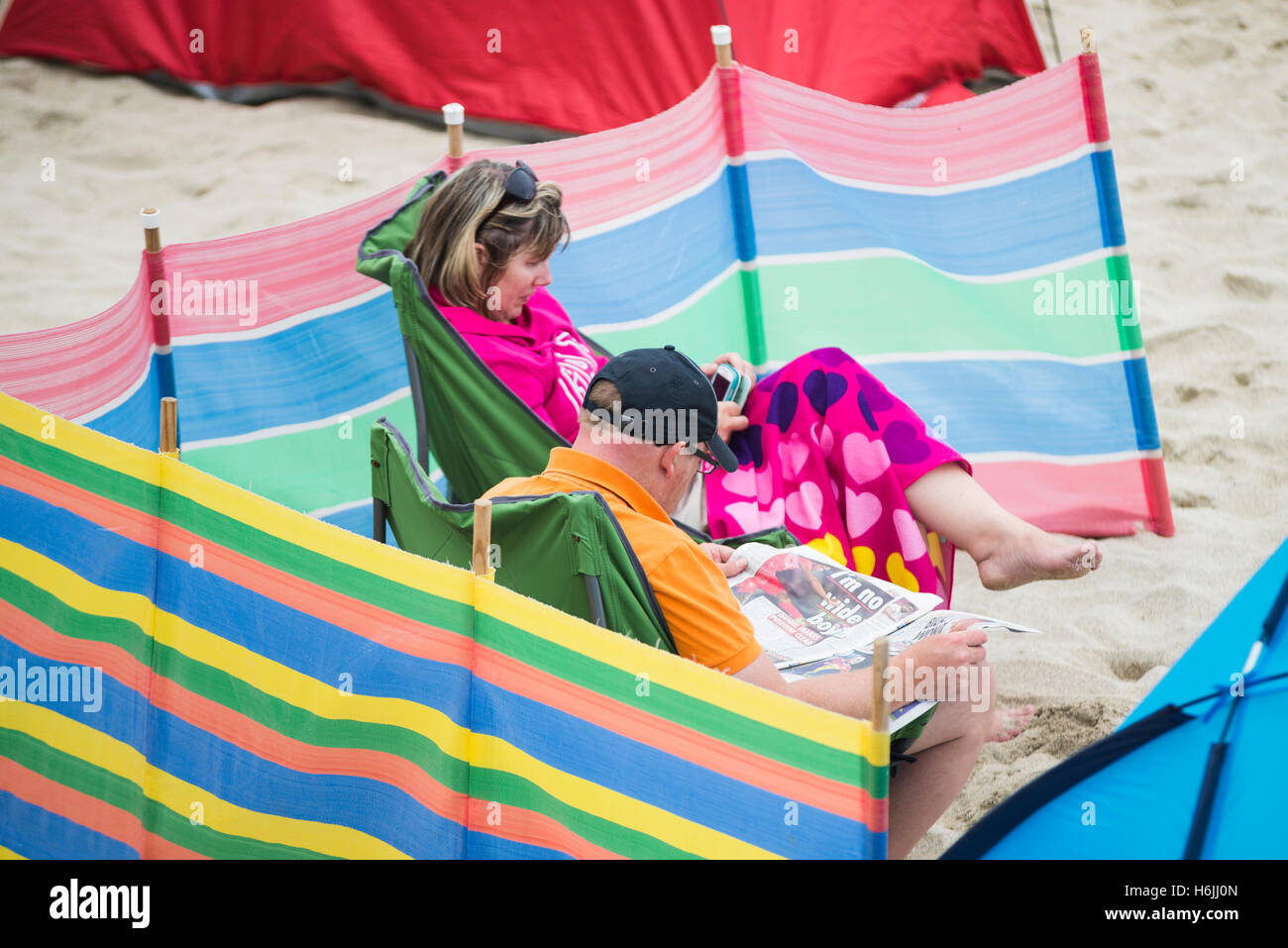 Ältere paar als Urlauber am Strand von St Ives sitzt und liest zwischen einem bunten Windschutz an einem bewölkt und windig Sommertag, Cornwall, UK Stockfoto