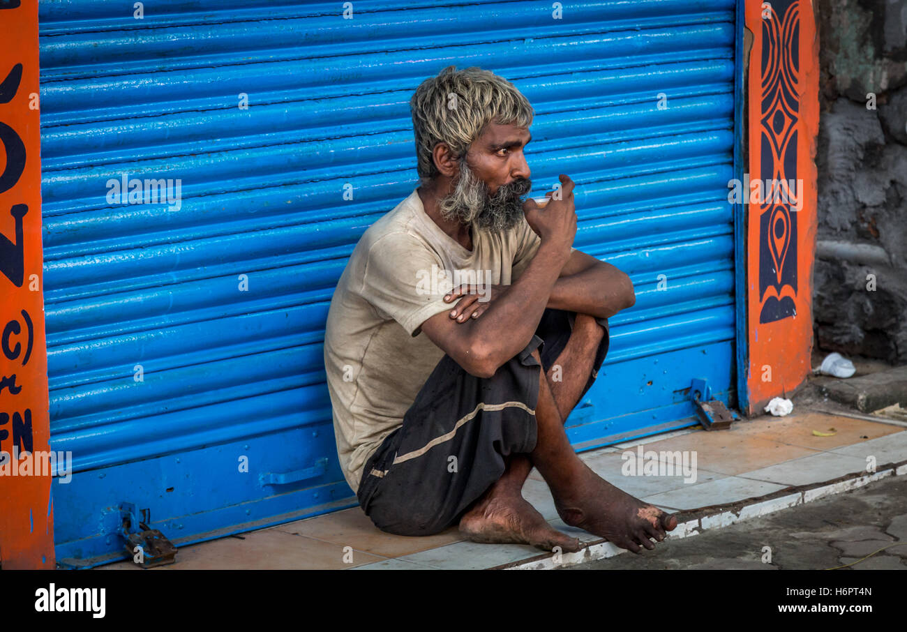 Alter indische Bettler sitzt vor einem Closed-Shop und hat eine Tasse Morgentee am auf einer Straße in South Kolkata, Westbengalen, Indien. Stockfoto