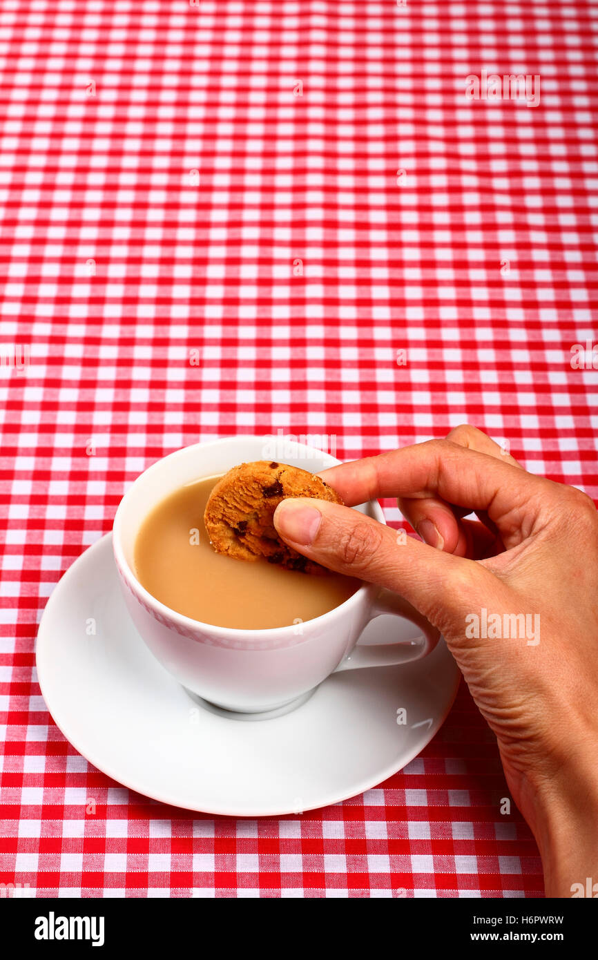 Frau-Damen-Girls Hand Eintunken einen Cookie Keks in zu einer heißen Tasse Tee mit einer roten und weißen aufgegebenes Tabelle Tuch Abdeckung Stockfoto
