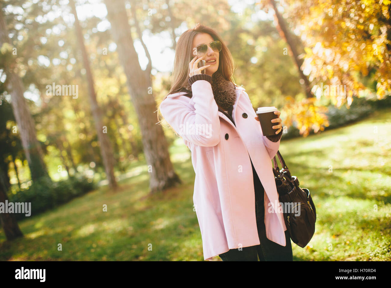 Junge Frau in den rosa Mantel im Gespräch über das Smartphone und hält Kaffee in der hand im Herbst Park gehen Stockfoto