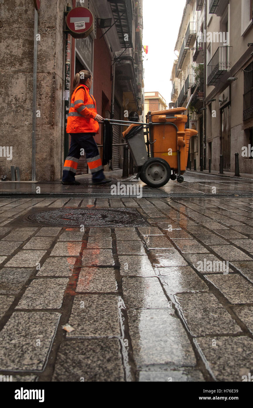 Frau Kehrmaschine mit orangefarbenen Arbeitskleidung, zu Fuß mit einem städtischen Reinigungswagen, durch die Straßen von Valencia, Spanien, mit dem Kopfsteinpflaster Boden nass Stockfoto