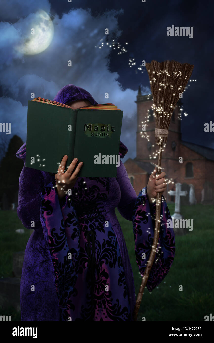 Frau trägt Purpurmantel im Friedhof mit Buch der Zaubersprüche und Besen Stockfoto