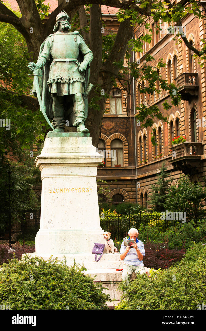 Ein Mann entspannt sich und liest sein Buch neben einer Statue an der Andrassy Avenue, während sein Hund Armbanduhr. Stockfoto