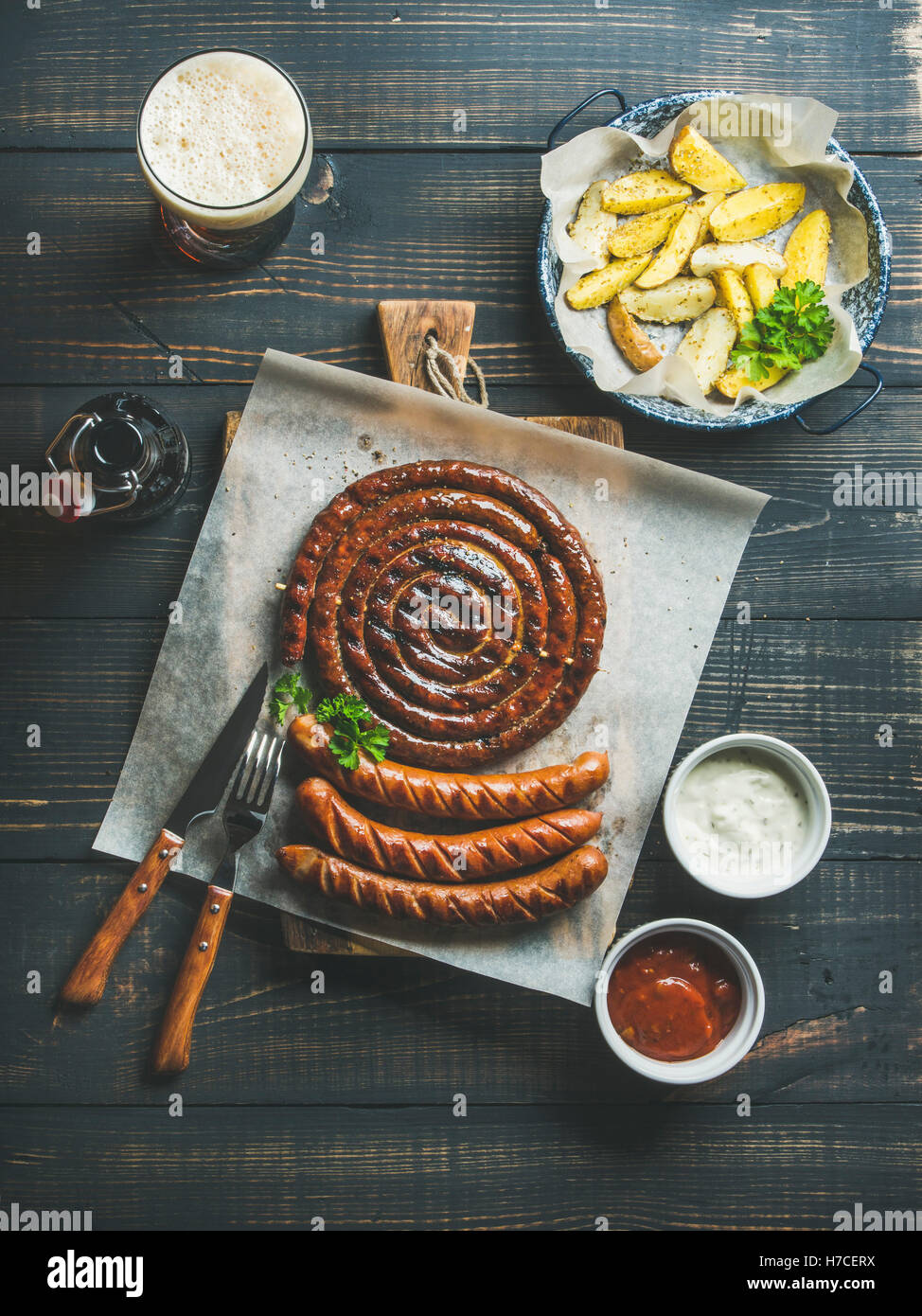 Gegrillte Würstchen mit Saucen, geröstete Kartoffel und Glas dunkles Bier auf rustikalen hölzernen Portion Brett über dunkle verbrannten Holz bac Stockfoto