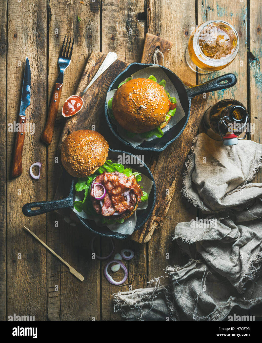 Hausgemachte Rindfleisch-Burger mit knusprigem Speck und Gemüse in kleine Gusseisen Pfannen und Glas Weißbier auf rustikalen Brett über shab Stockfoto