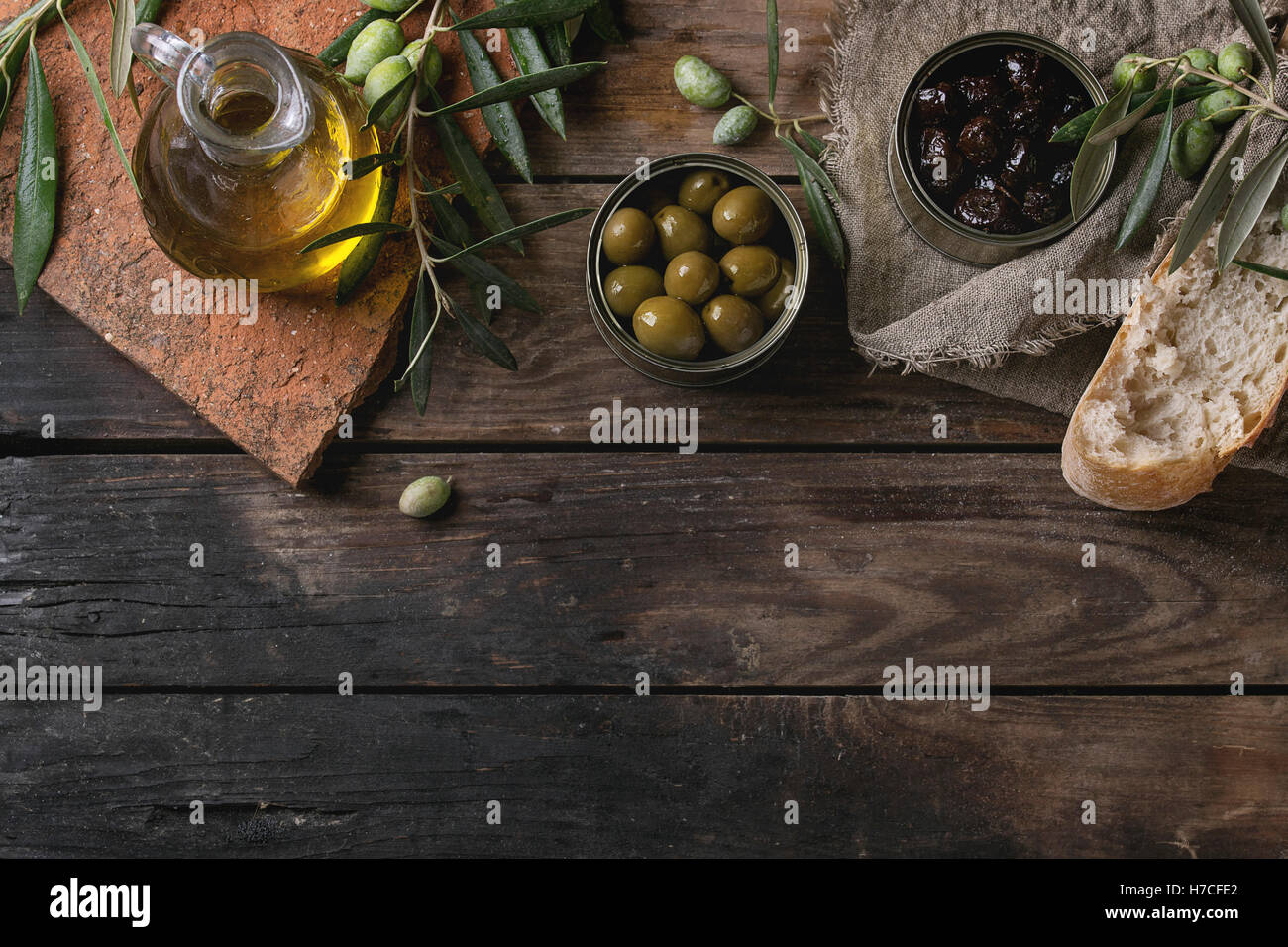 Grüne und schwarze Oliven in Blechdosen mit frischem Brot und junge Oliven verzweigen auf Sackleinen, Flasche Olivenöl auf Ton Bo Stockfoto
