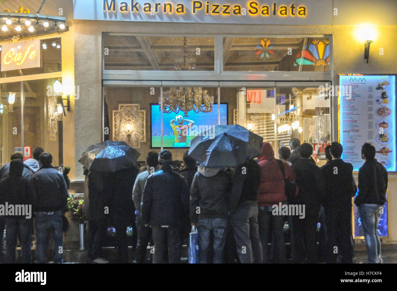 Menschen sehen ein Fußballspiel auf der großen Leinwand ein Restaurant, da sie draußen auf den Regen auf der Istiklal Straße in Istanbul stehen. Stockfoto