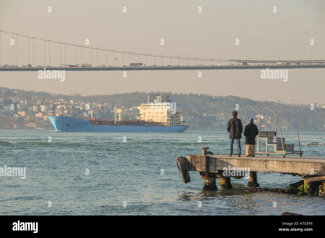 Die Menschen sehen eine Ladung Schiff Segel unter der Bosporus-Brücke aus einem Pier auf der asiatischen Seite. Stockfoto