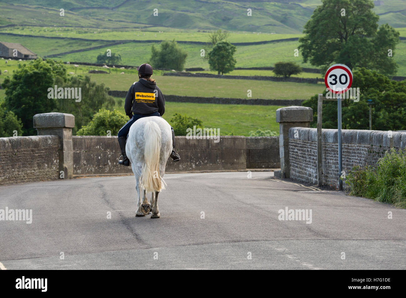 Rückansicht der jungen Frau, auf einem grauen Pferd auf einer ruhigen, malerischen, Country Road - Burnsall Dorf, Yorkshire Dales England. Stockfoto