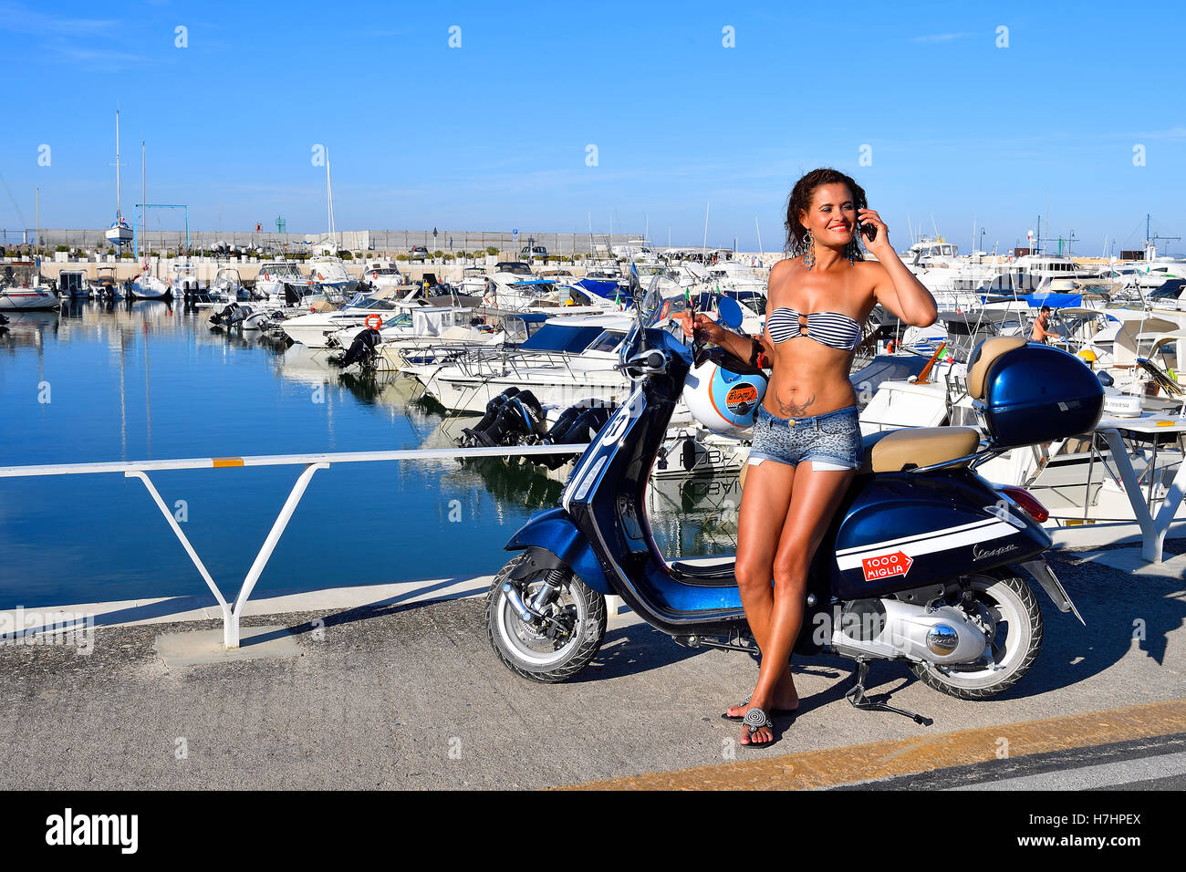Frau im Bikini-Oberteil mit Handy lehnt sich an eine Vespa Primavera Roller, Senigallia, Adria-Küste, Marche, Italien Stockfoto