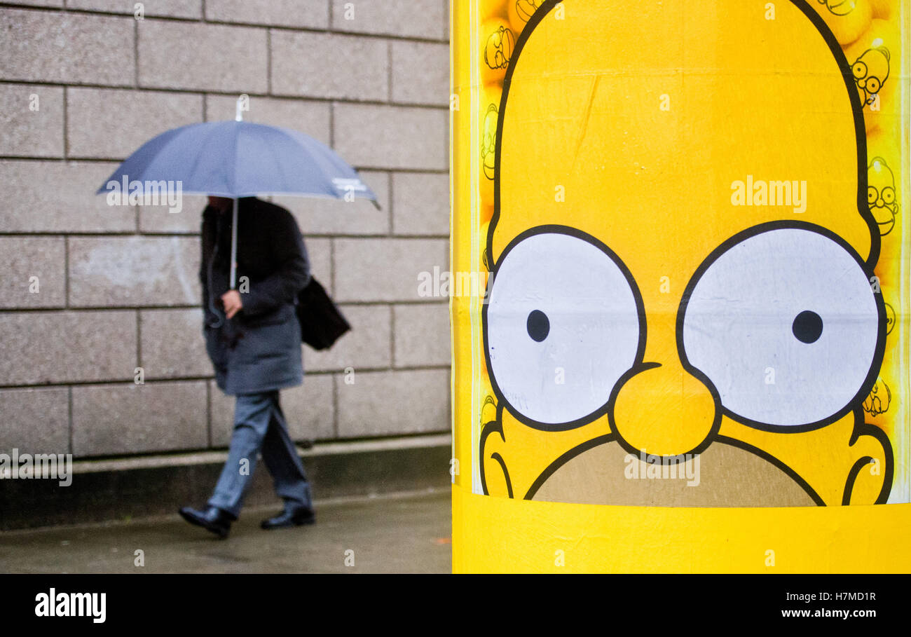 Hannover, Deutschland. 7. November 2016. Ein Mann mit einem Regenschirm  geht vorbei an einer Säule mit der Zeichentrickfigur Homer Simpson aus der  Zeichentrickserie Die Simpsons in Hannover, Deutschland, 7. November 2016.  Foto: