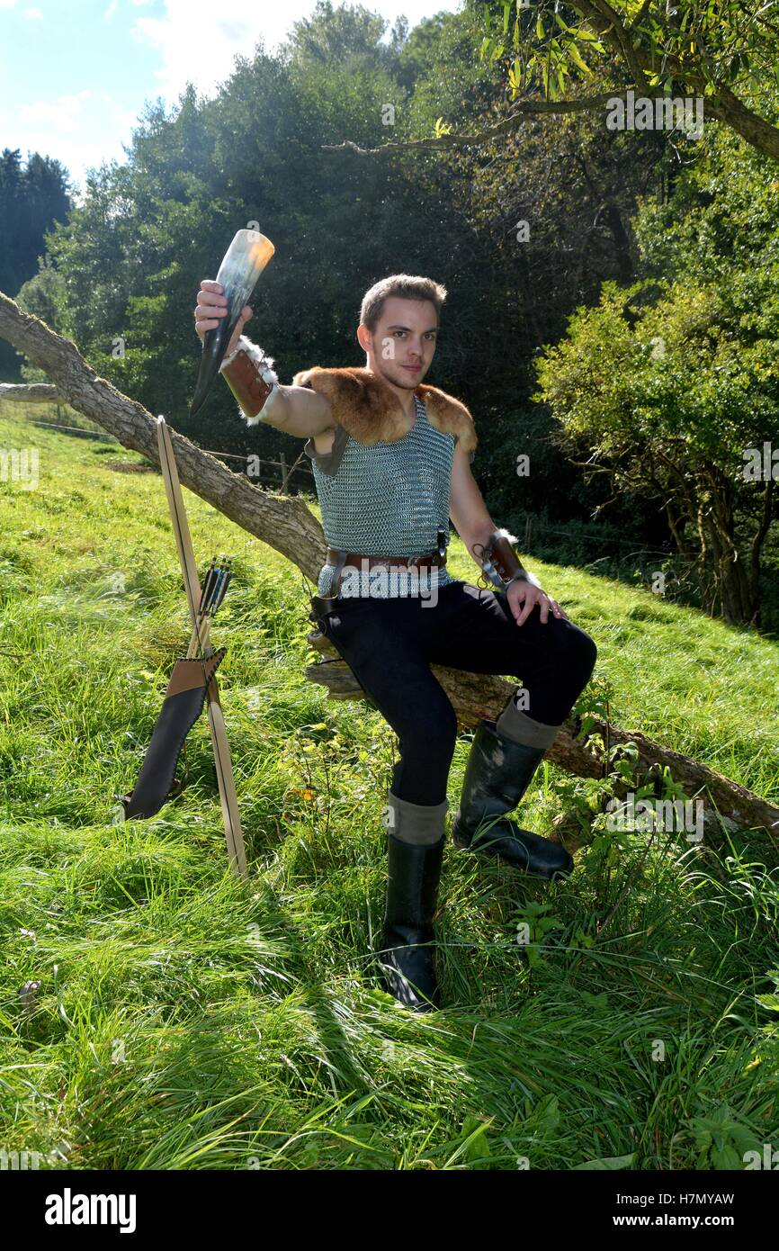 Mittelalterliche junge Mann, gekleidet hält Trinkhorn in der Hand und sitzt hoch auf Zweig in der Natur Stockfoto